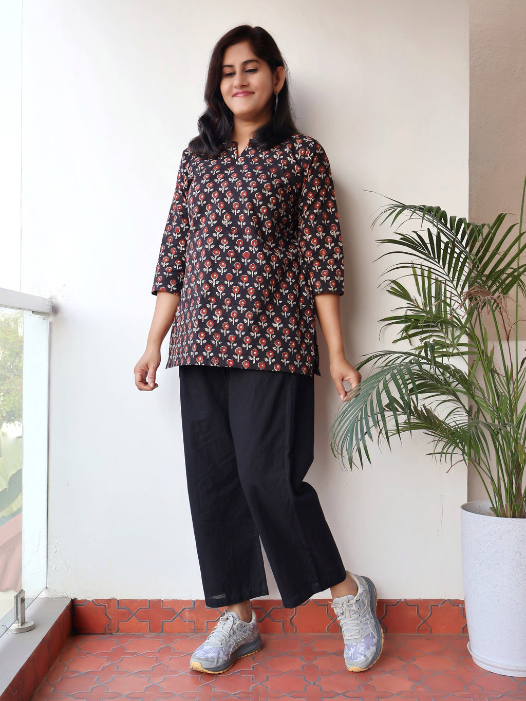 Woman wearing a patterned top and black pants standing indoors with a plant in the background