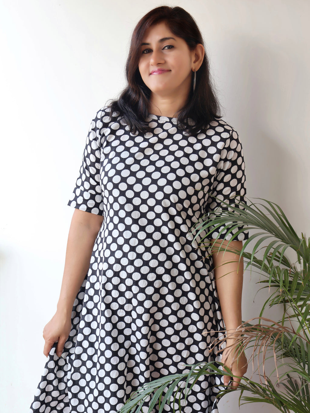 Woman wearing a black and white polka dot dress standing next to a plant against a plain background