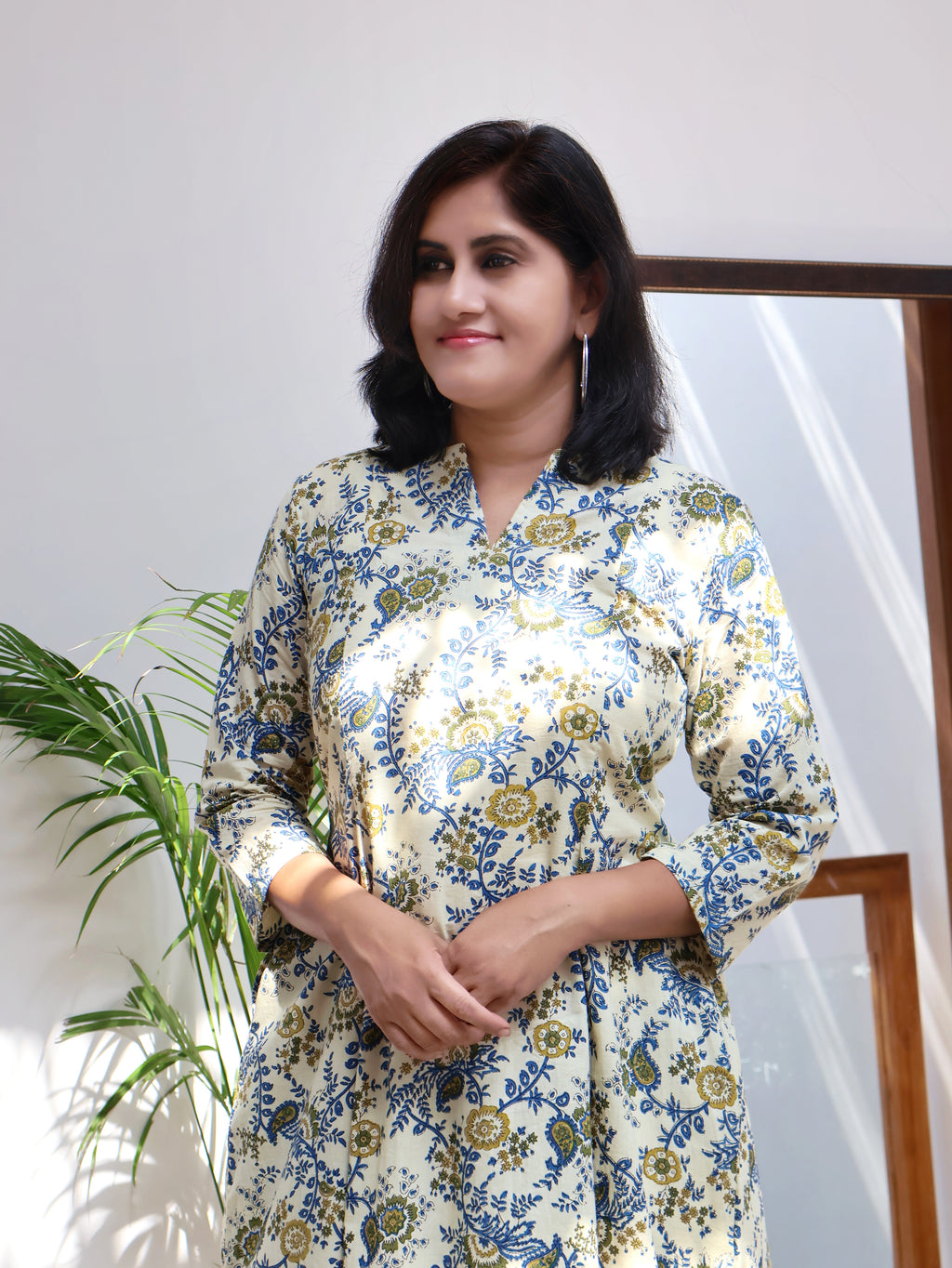 A person standing in a room with a plant to their left, wearing an off white and blue floral bagru hand block paneled long kurta with three-quarter sleeves.