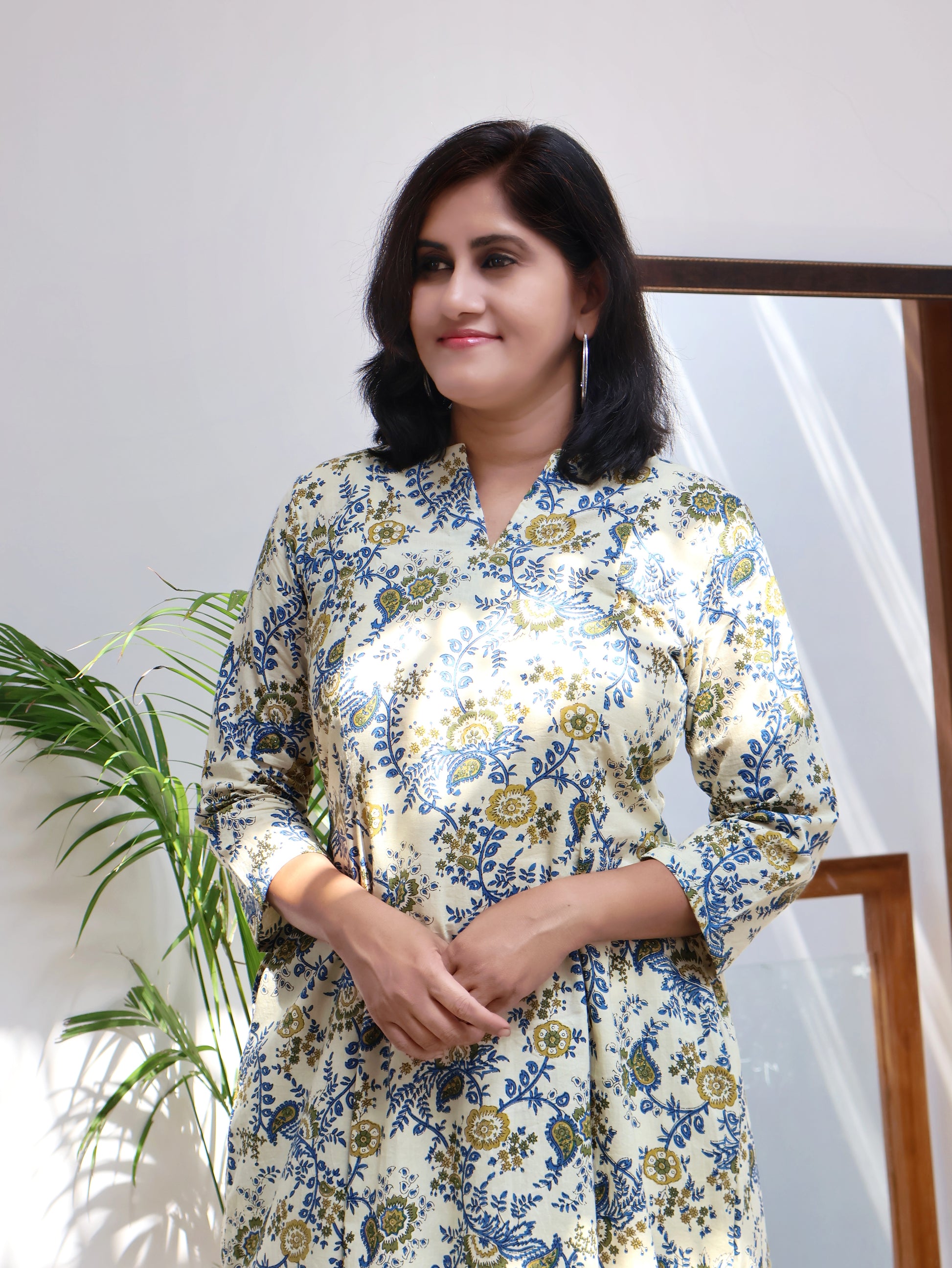A person standing in a room with a plant to their left, wearing an off white and blue floral bagru hand block paneled long kurta with three-quarter sleeves.