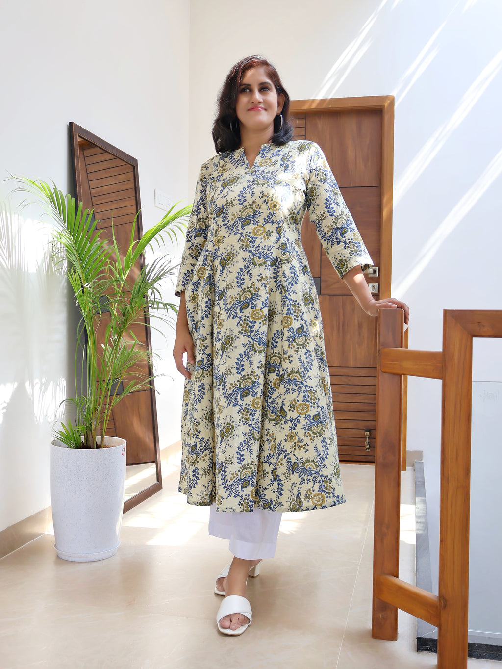 A woman standing in an indoor setting wearing an off white and blue floral long kurta with paneled design.