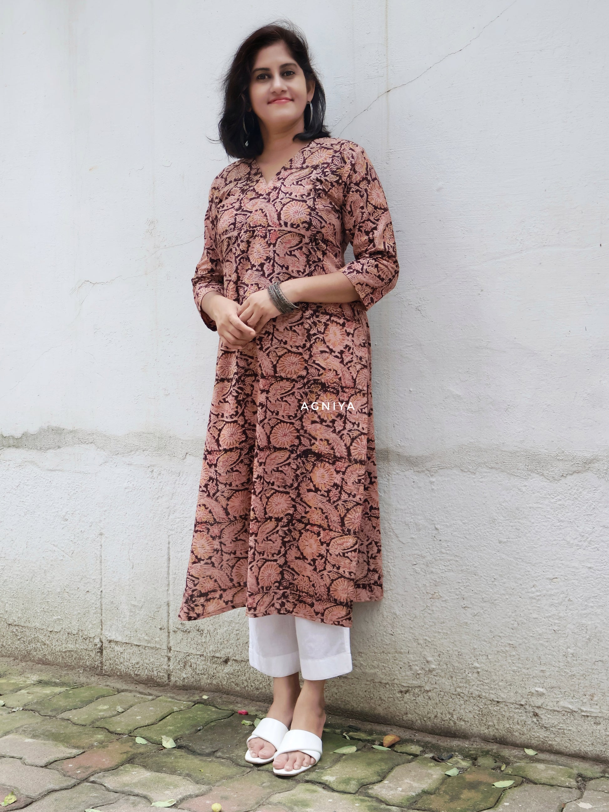 Woman wearing a patterned Kalamkari kurta and white pants standing against a white wall.
