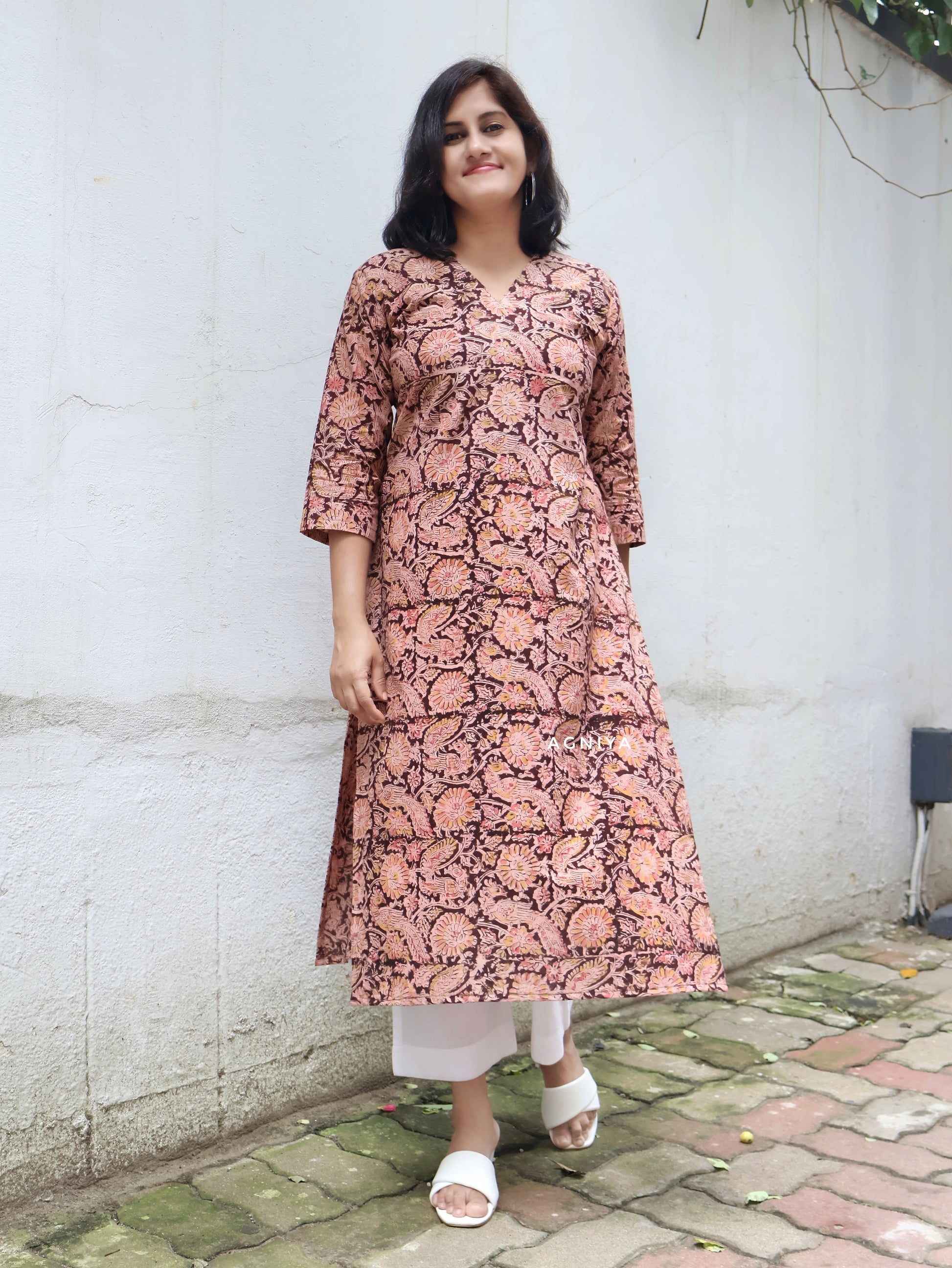 A woman standing on a footpath, wearing a maroon straight kurta with a floral hand block print, paired with white pants and white sandals.