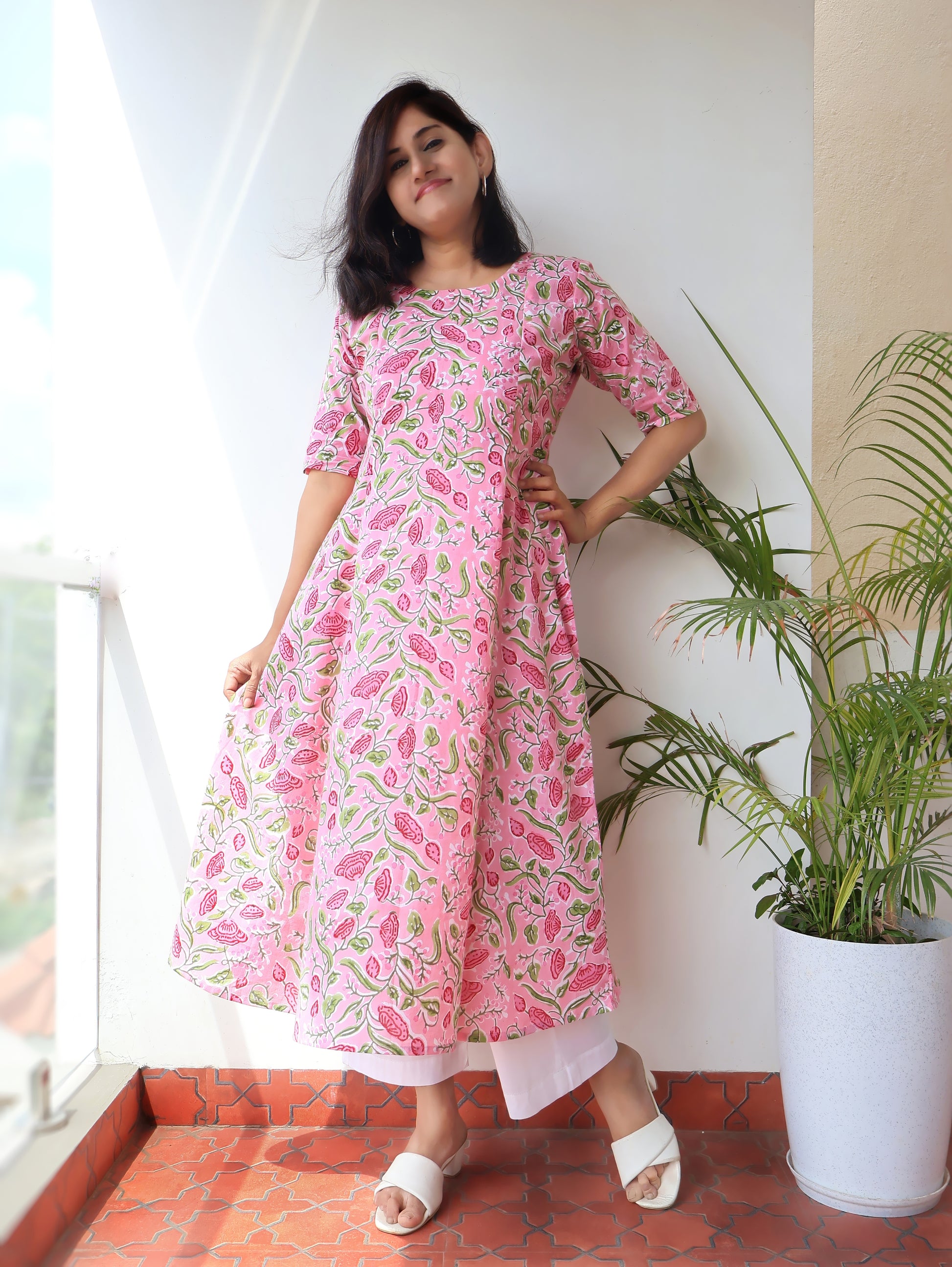 A woman modeling a pink and green floral print kurta with elbow-length sleeves, standing on a terracotta tiled balcony next to a potted plant.