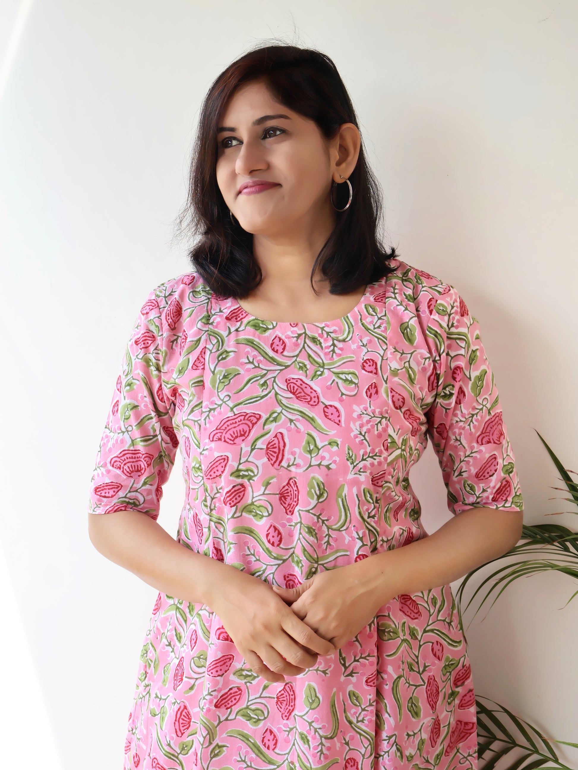 A woman wearing a pink cotton kurta with a green and red floral block print, standing against a plain white wall.