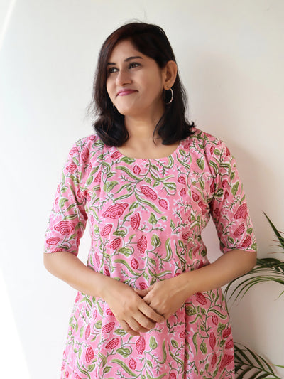 A woman wearing a pink cotton kurta with a green and red floral block print, standing against a plain white wall.